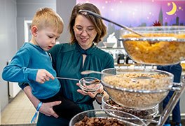 Mutter trägt ihren Sohn. Zusammen stehen sie vor der Müslibar in der Jugendherberge. Der Junge füllt Müsli in eine Schale, die die Mutter hält.