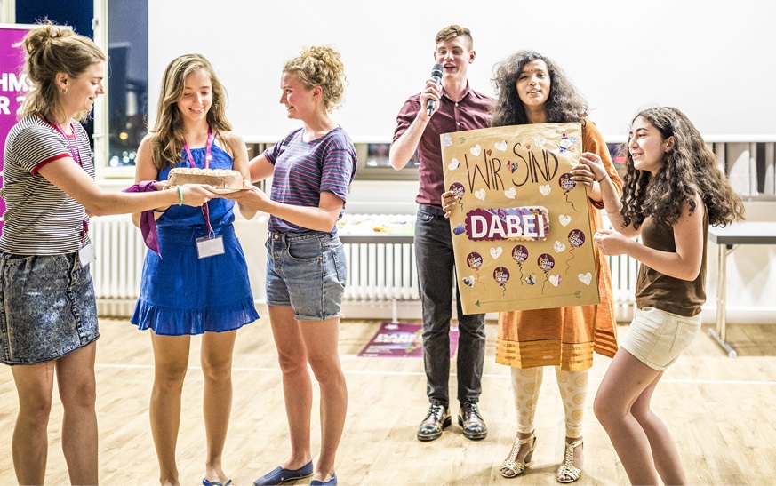 Gruppenveranstaltung in der Jugendherberge. Zwei Frauen überreichen einer anderen Frau im blauen Kleid einen Kuchen. Rechts halten zwei Mädchen ein Plakat hoch, auf dem steht „Wir sind dabei!“. Im Hintergrund steht ein Mann und spricht in ein Mikrophon.