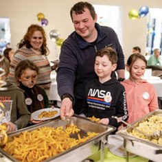 Mehrere Kinder stehen am Büfett in der Jugendherberge Trier. Ein Mann nimmt mit der Kelle Pommes.