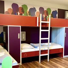 Ein Stockbett im Zimmer der Jugendherberge Mainz. Das Bett ist rot, die Schränke links und rechts sind lila. Die Wand zeigt viele bunte Silhouetten von Personen. 