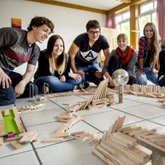 Fünf Personen sitzen im Halbkreis und beobachten gespannt, wie eine gebaute Reihe an Domino-Steinen umfällt.