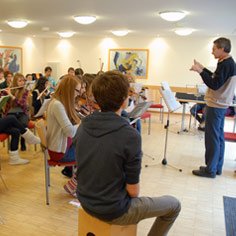 Schüler sitzen im Halbkreis und  proben auf Instrumenten in der Jugendherbergen Bad Marienberg. Der Dirigent steht rechts und gibt den Takt vor.